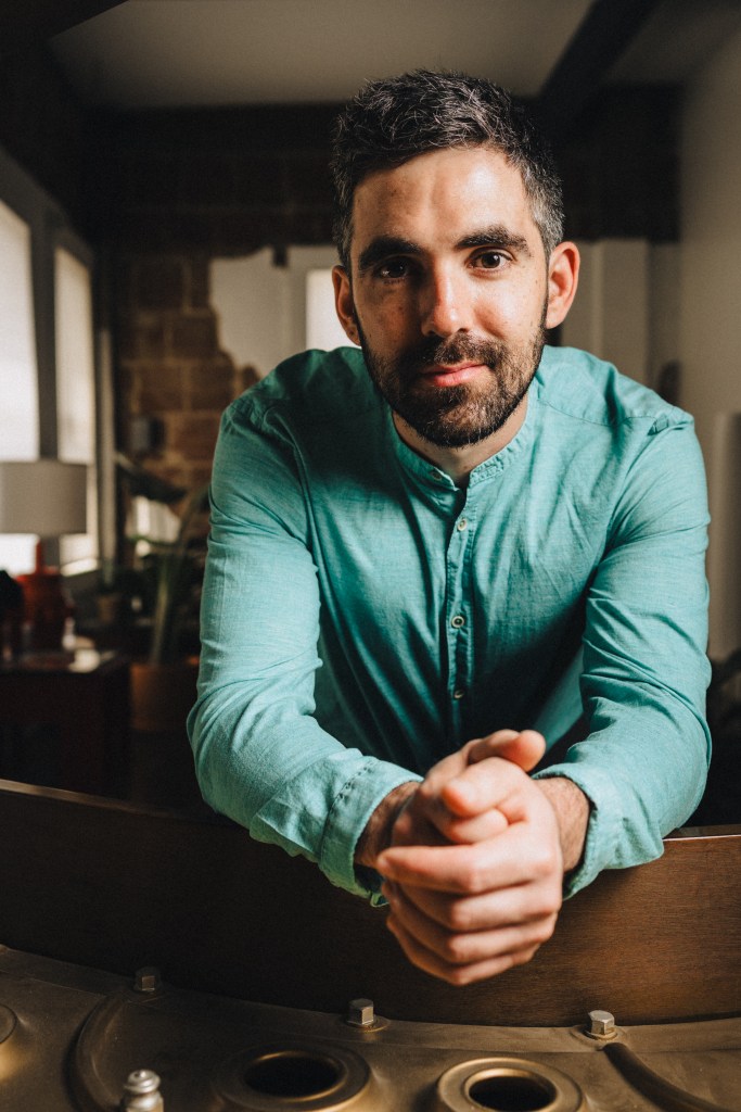 A person wearing a teal long-sleeve shirt leans forward over a wooden surface with their hands clasped together. They are indoors in a warmly lit room with large windows on the left and various furniture and decor in the background.